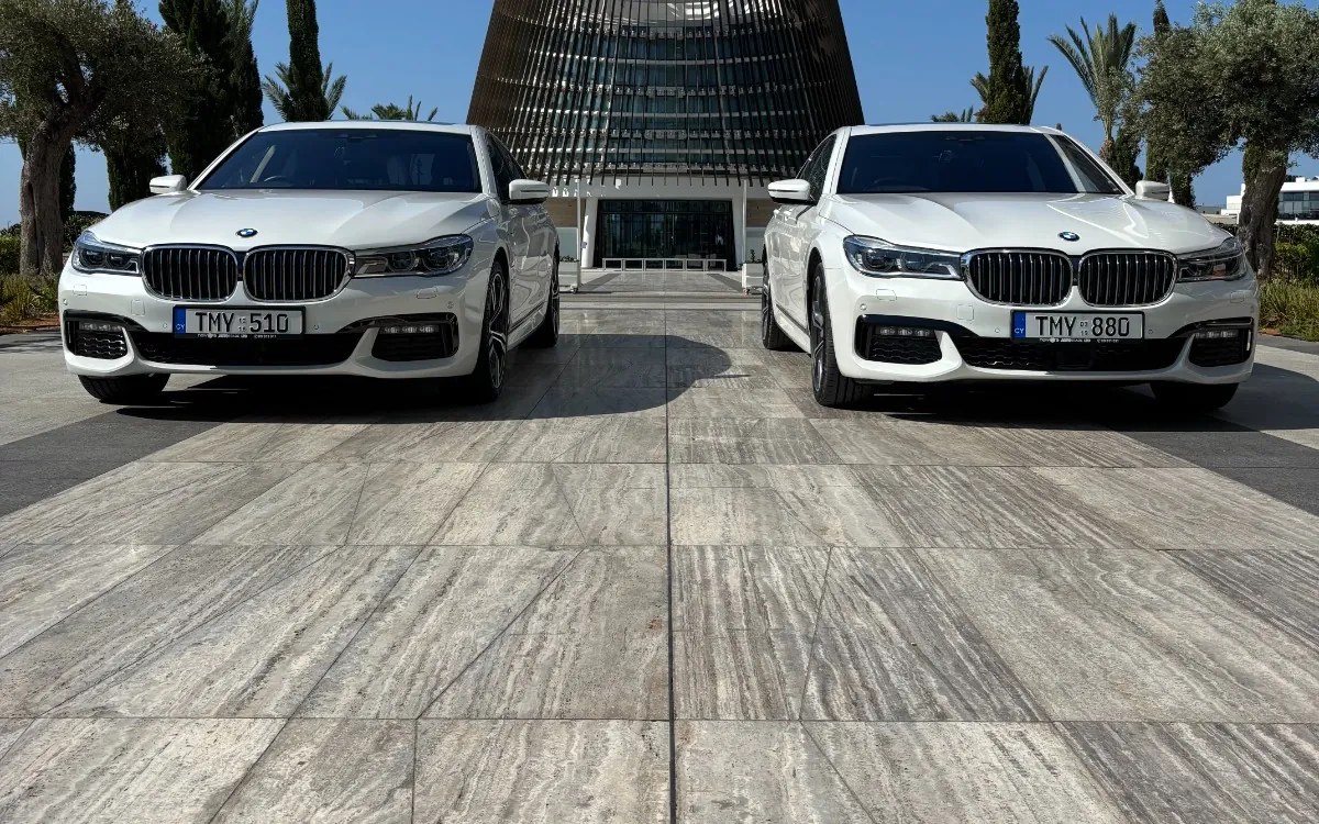 Two white BMW 7 Series sedans parked in front of a modern hotel in Cyprus